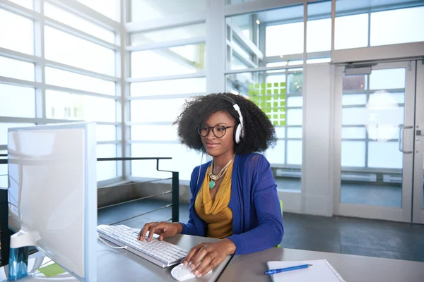 Retrato de uma mulher sorridente com um afro no computador em escritório de vidro brilhante — Fotografia de Stock