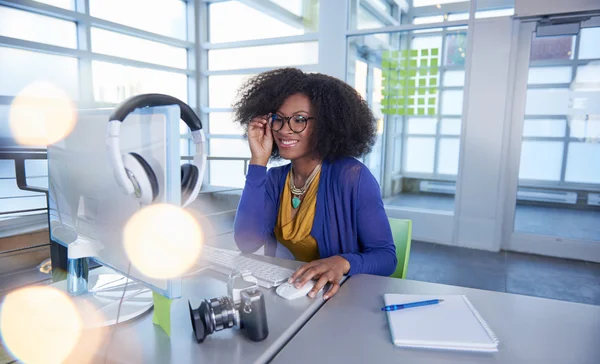 Portrait of a smiling woman with an afro at the computer in bright glass office — Stock Photo, Image