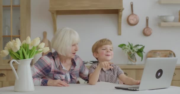 Grandmother and grandson talking online by laptop sitting at wooden kitchen. Smiling family having online conversation with relatives. Senior woman and kid watching cartoons on laptop. — Stock Video