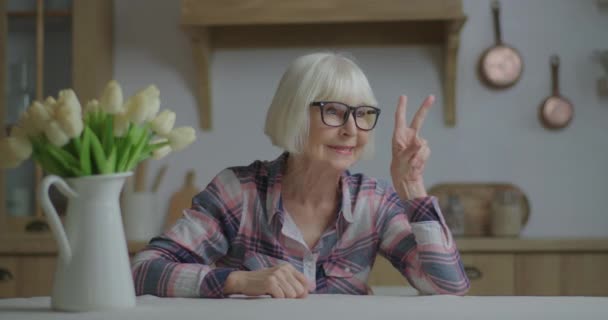 Смішна 70-ті жінка в окулярах показує знак перемоги пальцями, сидячи за обіднім столом з білими тюльпанами, дивлячись на камеру. Щаслива старша блондинка показує V знак і язик на камеру і посміхається . — стокове відео