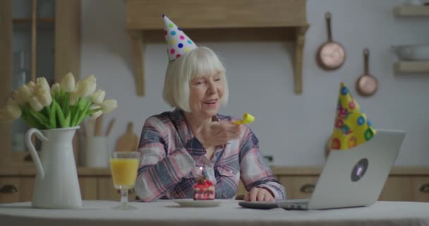 Χαρούμενη ηλικιωμένη γυναίκα γιορτάζει τα γενέθλιά της στο διαδίκτυο με φορητό υπολογιστή. 70άρα γυναίκα με καπέλο γενεθλίων να φυσάει πίπα γενεθλίων σε φίλους και συγγενείς μέσω διαδικτύου. Κοινωνική απόσταση και χαιρετισμός μέσω βιντεοκλήσης. — Αρχείο Βίντεο