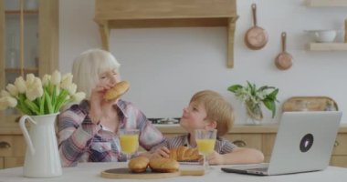 Büyükannem ve torunum çörek yer ve portakal suyu içerler. Mutfaktaki yemek masasında otururlar. Aile ev yapımı pastayı sever..