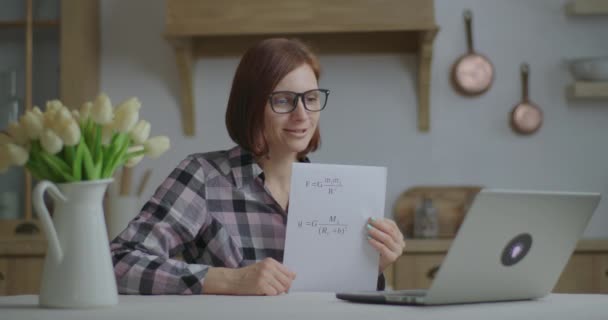 Młoda 30-letnia nauczycielka w okularach rozmawia i pokazuje kartkę papieru z równaniami fizyki patrząc na laptopa w kuchni. Edukacja online i proces uczenia się na odległość. Praca nauczyciela w domu. — Wideo stockowe