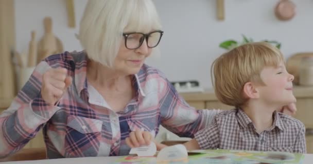 Vorschulkind lacht und spielt Brettspiel mit Großmutter in Holzküche. Glückliche Familie beim gemeinsamen Lesen interessanter Bücher. Oma und Enkelkind haben Spaß. — Stockvideo