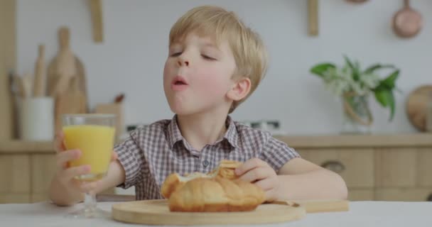 Förskolekillen äter bullar och dricker apelsinjuice i träköket. Hungrig unge tuggar hembakat bakverk hemma. — Stockvideo