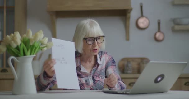 Profesora sénior en gafas habla y muestra hoja de papel con ecuaciones físicas mirando el portátil en la cocina. Educación en línea y proceso de aprendizaje a distancia. Trabajo del profesor en casa. — Vídeo de stock