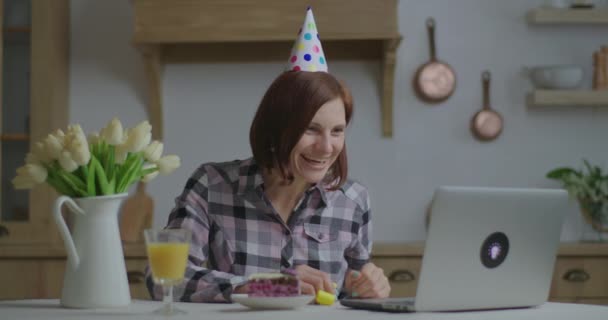 Wanita berusia 30-an dengan topi ulang tahun berbicara online dengan laptop. Happy person merayakan ulang tahun secara online di rumah sendirian dengan sepotong kue dan jus jeruk. Jarak sosial dan salam melalui panggilan video. — Stok Video