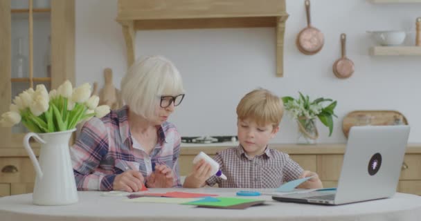 Aile evde başvuru yapıyor. Büyük anne ve torunu mutfakta yapıştırıcı, makas ve renkli kağıtlarla yaratıcı aktiviteler yapıyorlar. Birlikte sanat yapmak.. — Stok video