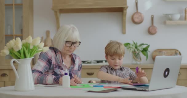 Anaokulu öğrencisi yapıştırıcı, makas ve renkli kağıtla resim ödevi yapıyor ve çizimlerini laptopa gösteriyor. Büyük büyükanne çocuğa yardım ediyor. Aile birlikte sanat yapıyor. Çevrimiçi eğitim süreci. — Stok video