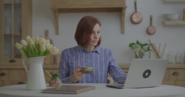 Joven mujer de negocios de 30 años escribiendo en el ordenador portátil y beber café en la cocina. Empresaria cansada de trabajar online desde casa. Mujer cierra portátil y disfruta del café. — Vídeos de Stock