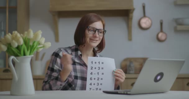 Jonge dertiger jaren vrouwelijke leraar in bril praat en toont vel papier met eenvoudige wiskundige vergelijkingen op zoek naar laptop. Online onderwijs en afstandsonderwijs. Werk van thuis wiskundeleraar. — Stockvideo