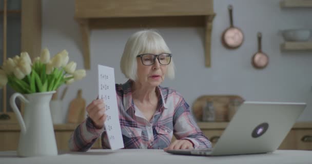 Profesora sénior en gafas habla y muestra hoja de papel con ecuaciones matemáticas simples mirando el portátil en la cocina. Educación en línea y proceso de aprendizaje a distancia. Trabajo del profesor en casa. — Vídeos de Stock