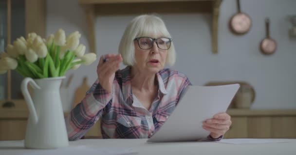 Χαμογελώντας ηλικιωμένη γυναίκα με γυαλιά διαβάζει κείμενο σε χαρτί δυνατά και κάνει σημειώσεις με στυλό. Εκπαιδευτική διαδικασία. Εργασία από το σπίτι γυναίκα μιλώντας κατά την ανάγνωση. — Αρχείο Βίντεο