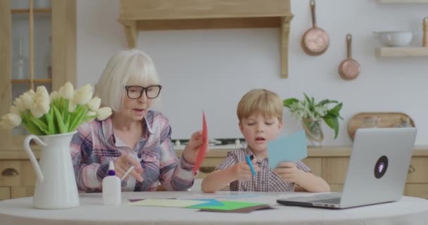 Starší babička se přihlásila k vnukovi ze školky doma. Rodina dělá tvůrčí činnost s lepidlem, nůžkami a barevným papírem v kuchyni. Tvorba umění společně. — Stock video