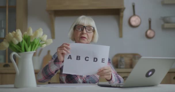 Eine Lehrerin mit Brille spricht und zeigt ein Blatt Papier mit Buchstaben aus dem Alphabet, das in der Küche auf den Laptop schaut. Online-Bildung und Fernunterricht. Arbeit von der Hauslehrerin. — Stockvideo