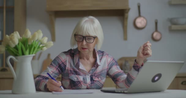 ノートパソコンを見て紙にペンで書く眼鏡の上級教師。オンライン教育と距離学習プロセス。自宅の女性の署名文書から働く. — ストック動画