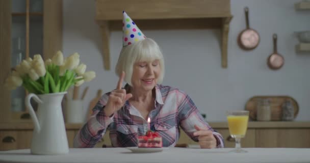 Fröhliche Seniorin feiert Geburtstag allein mit Torte und Kerze. 70er Jahre Frau mit Geburtstagshut bläst Geburtstagskerzen an und bleibt allein zu Hause. — Stockvideo