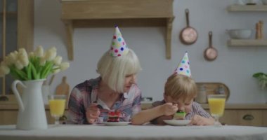 Mutlu büyükanne torunuyla birlikte evde doğum gününü kutluyor. Doğum günü şapkalı aile, ahşap mutfakta doğum günü pastası yerken eğleniyor..