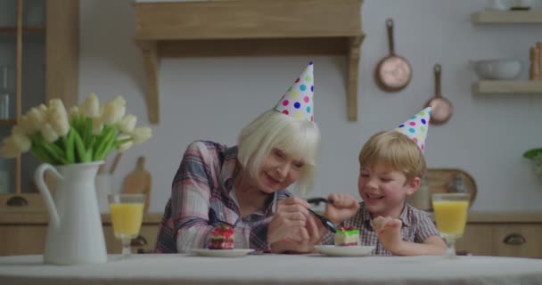 Vrolijke oudere vrouw viert verjaardag met kleinzoon samen thuis. Familie in verjaardagshoeden hebben plezier tijdens het eten van verjaardagstaart in houten keuken. — Stockvideo