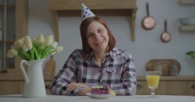 Doğum günü şapkalı genç bir kadın kameraya bakarak konuşuyor. Mutlu insan doğum gününü internette tek başına pasta ve portakal suyuyla kutlar. Video çağrısı yoluyla doğum günü iletisinin webcam görünümü.