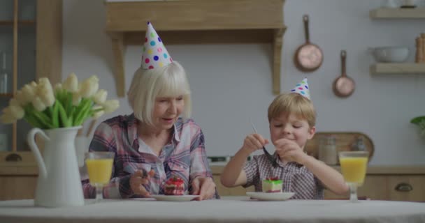 Веселая бабушка празднует день рождения вместе с внуком дома. Семья в шляпах на день рождения веселится, поедая торты на деревянной кухне. — стоковое видео