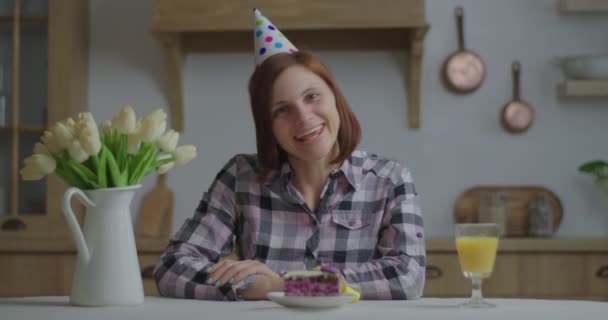 Młoda 30-latka w urodzinowym kapeluszu rozmawiająca przed kamerą. Happy person świętuje urodziny online sam z kawałkiem ciasta i soku pomarańczowego. Kamera internetowa widok urodzin powitanie przez rozmowy wideo. — Wideo stockowe