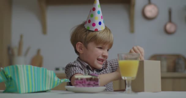 Счастливый ребенок открывает подарок на день рождения, сидя дома за столом. Радостный мальчик в шляпе на день рождения разворачивает подарок. — стоковое видео