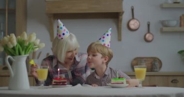 Parti şapkalı anaokulu çocuğu büyükannesinin doğum gününü evde kutluyor. Aile, doğum günü mumlarını bir parça kekin üzerine üflüyor. Evde doğum günü partisi.