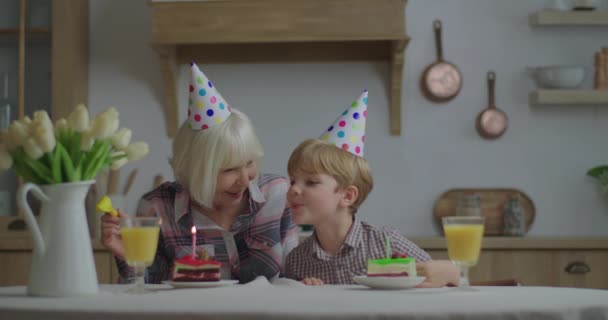 Parti şapkalı anaokulu çocuğu büyükannesinin doğum gününü evde kutluyor. Aile, doğum günü mumlarını bir parça kekin üzerine üflüyor. Evde doğum günü partisi. — Stok video