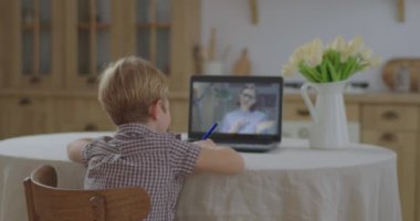Anaokulu çocuğu dizüstü bilgisayar ekranında bayan öğretmenle online okul ödevi yapıyor. Çocuk evde online çalışıyor. Çevrimiçi eğitim ve uzaktan öğrenme. Çocuk alıştırma kitabını çevrimiçi öğretmene gösterir. 
