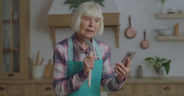 Femme blonde âgée joyeuse chantant dans les ustensiles de cuisine comme un microphone regardant le téléphone portable dans les mains à la cuisine en bois. Femme âgée qui s'amuse à rester à la maison. Scoop comme microphone. — Video