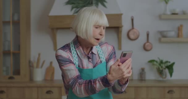 Vrolijke oudere blonde vrouw zingen naar mobiele telefoon in de handen van houten keuken. Oudere vrouw heeft plezier thuis te blijven. Grootmoeder met mobiele telefoon. — Stockvideo