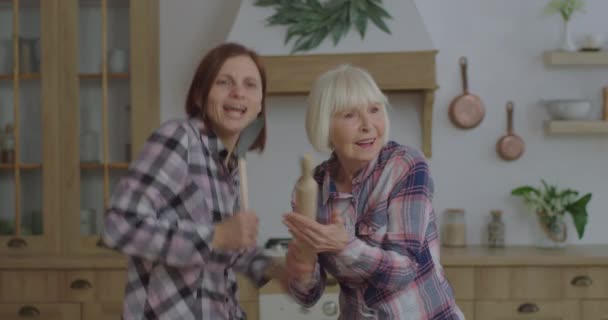 Lycklig familj av ung vuxen kvinna och äldre mor dansar och sjunger i köksutrustning som en mikrofon i köket. Kvinnor har kul med rullande stift och scoop vistas hemma. — Stockvideo