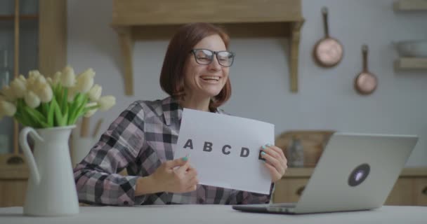 Unga 30-tals kvinnliga lärare i glasögon talar och visar pappersark med alfabet bokstäver tittar på laptop i köket. Utbildning på nätet och distansundervisning. Arbete från hemlärare. — Stockvideo