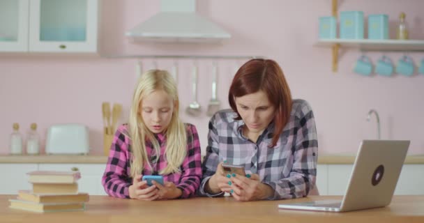 Szőke iskoláslány és fiatal anya böngészi az internetet mobiltelefonnal otthon ülve a rózsaszín konyhában. Anya és lánya együtt szórakoznak a kütyükkel. Lány néz online video a szociális hálózat. — Stock videók