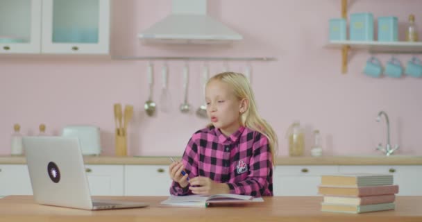La colegiala tiene clase en línea en casa. Niño hablando con profesor en línea mientras estudia en línea en el ordenador portátil. Chica de camisa rosa en la cocina rosa. — Vídeo de stock