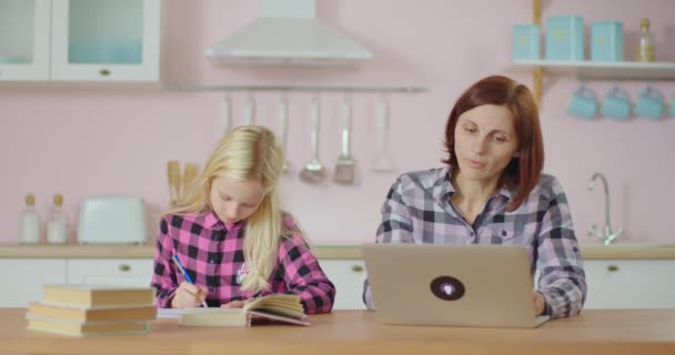 Kleuterschoolmeisje maakt huiswerk en moeder typt op laptop. Familie blijft samen thuis. Werken vanuit huis moeder met kind zitten in roze keuken. — Stockvideo