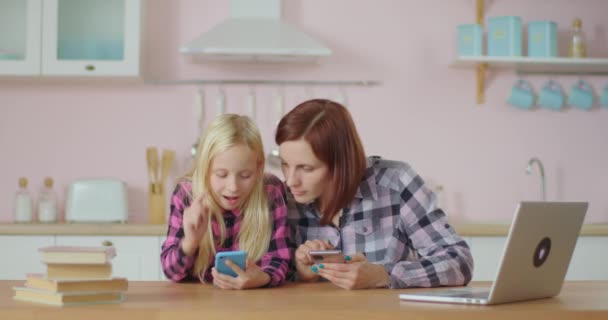 Блондинка школьница и молодая мать просматривают интернет по мобильным телефонам, сидя дома на розовой кухне. Мама и дочь вместе веселятся с гаджетами. Девушка смотрит онлайн видео в социальной сети. — стоковое видео