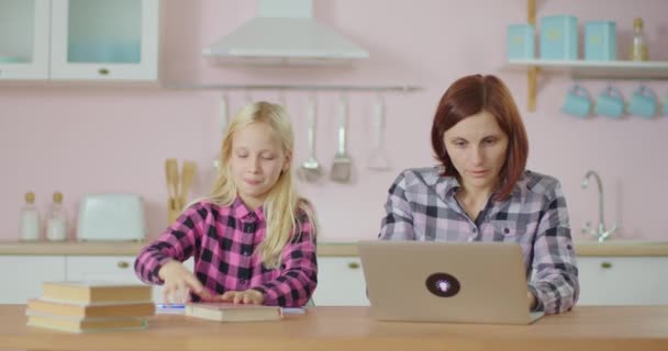 Menina da escola pré-adolescente fazendo lição de casa e mãe digitando no laptop. A família fica em casa juntos. Trabalhando a partir de casa mãe com criança sentada na cozinha rosa. — Vídeo de Stock