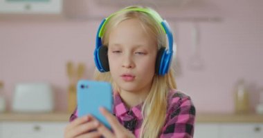 Renkli kulaklıklı, elinde cep telefonuyla pembe mutfakta oturan konsantre okul kızı. Genç kız cep telefonuna odaklanmış..