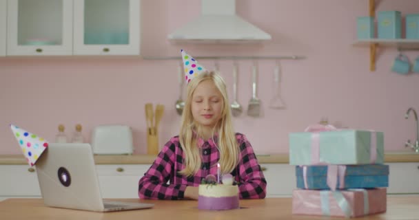 パーティーの帽子の誕生日の女の子は目を閉じて願いを作り、誕生日ケーキにキャンドルを吹く準備ができています。子供は孤独の中で一人誕生日を祝う. — ストック動画