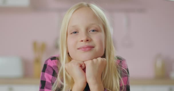 Портрет блондинки с длинными волосами школьницы, улыбающейся и смеющейся, глядя в камеру. Позитивный ребенок в розовой рубашке на розовой кухне. Закрыть счастливого малыша. — стоковое видео