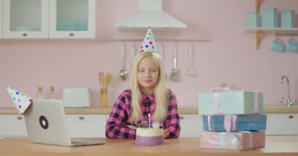 Η εορτάζουσα με το καπέλο κάνει μια ευχή με κλειστά μάτια και φυσάει κερί στην τούρτα γενεθλίων. Παιδί γιορτάζει μόνος γενέθλια με φίλους σε απευθείας σύνδεση κατά τη διάρκεια της απομόνωσης. — Αρχείο Βίντεο