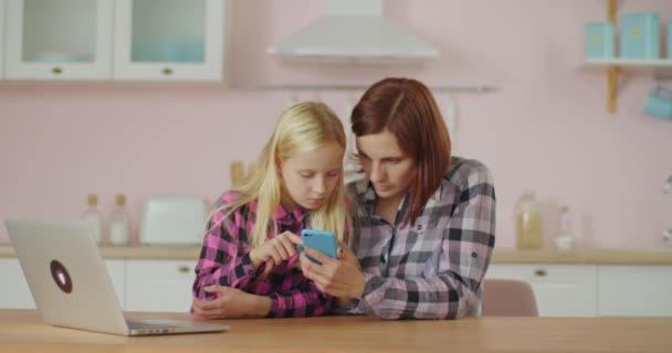 Ung 30-talskvinna surfar internet med skolflicka sitter hemma rosa kök. Mamma och dotter tittar på videor på mobiltelefon. — Stockvideo