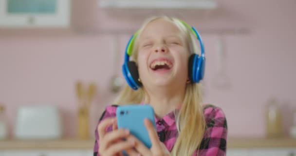 Niña de la escuela riendo en auriculares de color con teléfono móvil en las manos. Retrato de chica rubia divertida se ríe en casa. — Vídeos de Stock