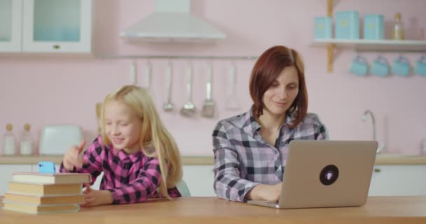 Jonge schoolmeisje opnemen video blog op mobiele telefoon met moeder werkt op laptop in de buurt. glimlachende vrouw geniet van tijd met dochter. — Stockvideo