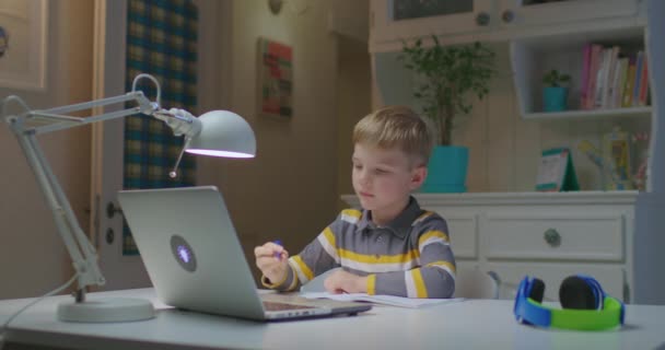 Niño preescolar mostrando cuaderno a la pantalla del ordenador portátil mientras estudia en línea en casa. Educación en línea y aprendizaje a distancia para niños. Niño haciendo la tarea por Internet. — Vídeos de Stock