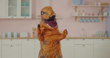 Kahverengi dinozorun pembe mutfakta komik dansı. Dinozor maskotunun komik hareketleri.