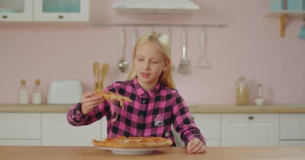 Χαριτωμένο κορίτσι με ροζ πουκάμισο που τρώει πίτσα στη ροζ κουζίνα. Στην μαθήτρια αρέσει να τρώει πίτσα με τα χέρια. Πεινασμένο παιδί στο σπίτι. — Αρχείο Βίντεο