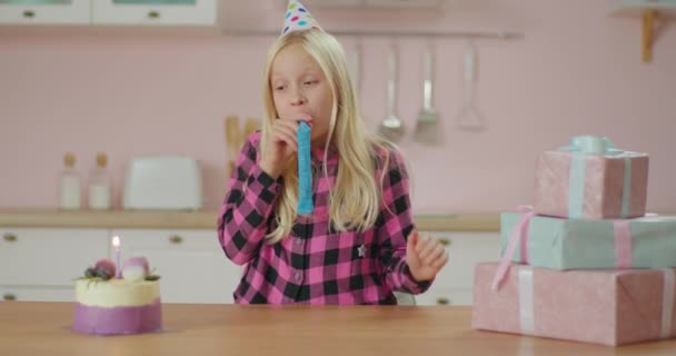 Досить маленька дівчинка святкує день народження, сидячи вдома наодинці. День народження дівчина робить бажання і дме свічку на торт з партійним свистом . — стокове відео